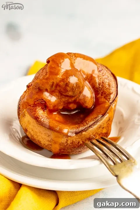 A Yorkshire pudding with gravy poured over it in a small white bowl set over a mustard yellow napkin. There's a fork resting on the bowl.