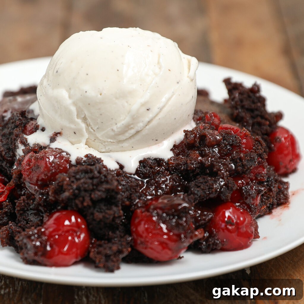 A serving of black forest cobbler topped with a scoop of vanilla ice cream on a white plate, placed on a rustic wooden surface.