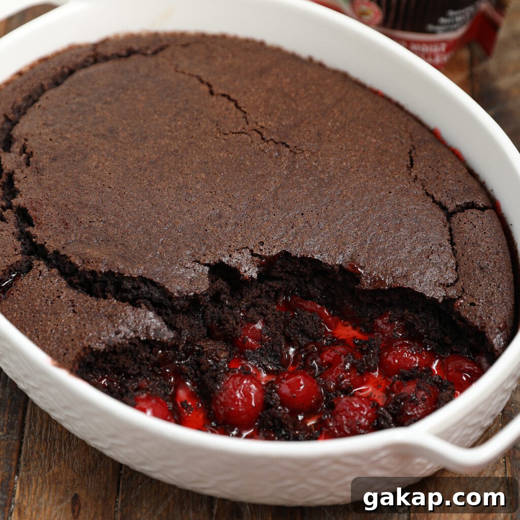 A baked black forest cobbler in a white oval dish, with a portion scooped out to reveal juicy red cherries and gooey chocolate beneath a cracked, cake-like top.