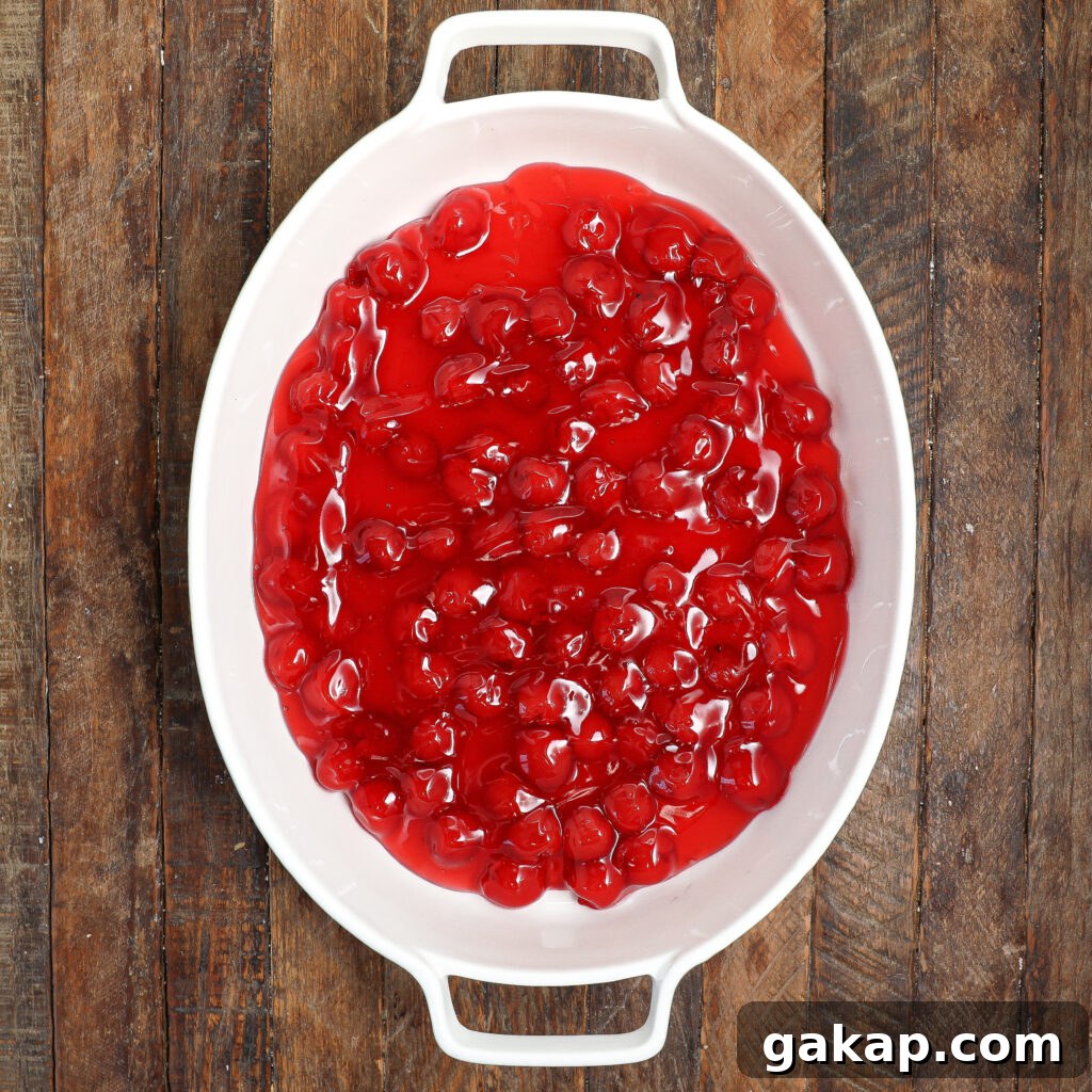 A white oval baking dish filled with vibrant black forest cobbler, featuring bright red cherry pie filling, sits on a rustic wooden table.