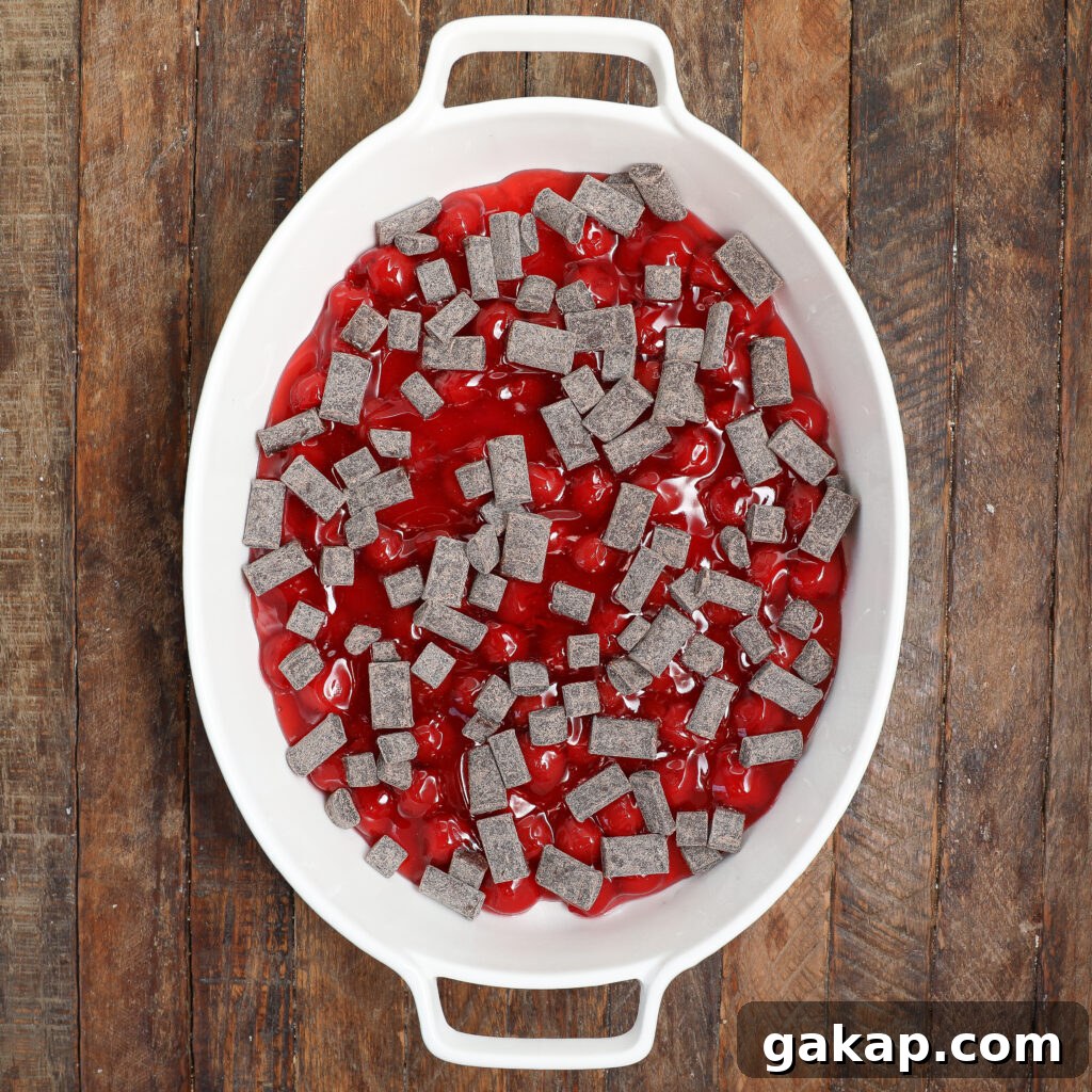 A white oval baking dish filled with cherry pie filling and topped with rectangular pieces of chocolate, creating a delicious black forest cobbler, is placed on a rustic wooden surface.