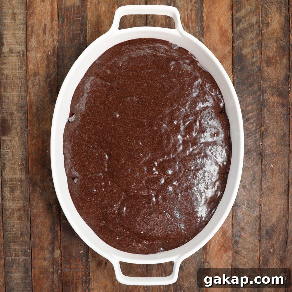 Oval white baking dish filled with smooth, uncooked black forest cobbler chocolate batter, placed on a rustic wooden surface.