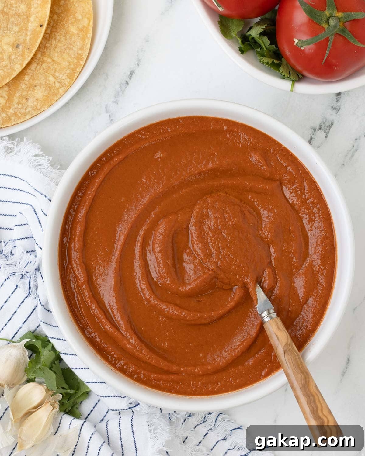 A bowl of deep red ranchero sauce in a white bowl with spoon, garlic cloves, and tortillas.